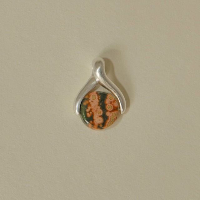 Silver pendant with a circular green and orange stone on a beige background